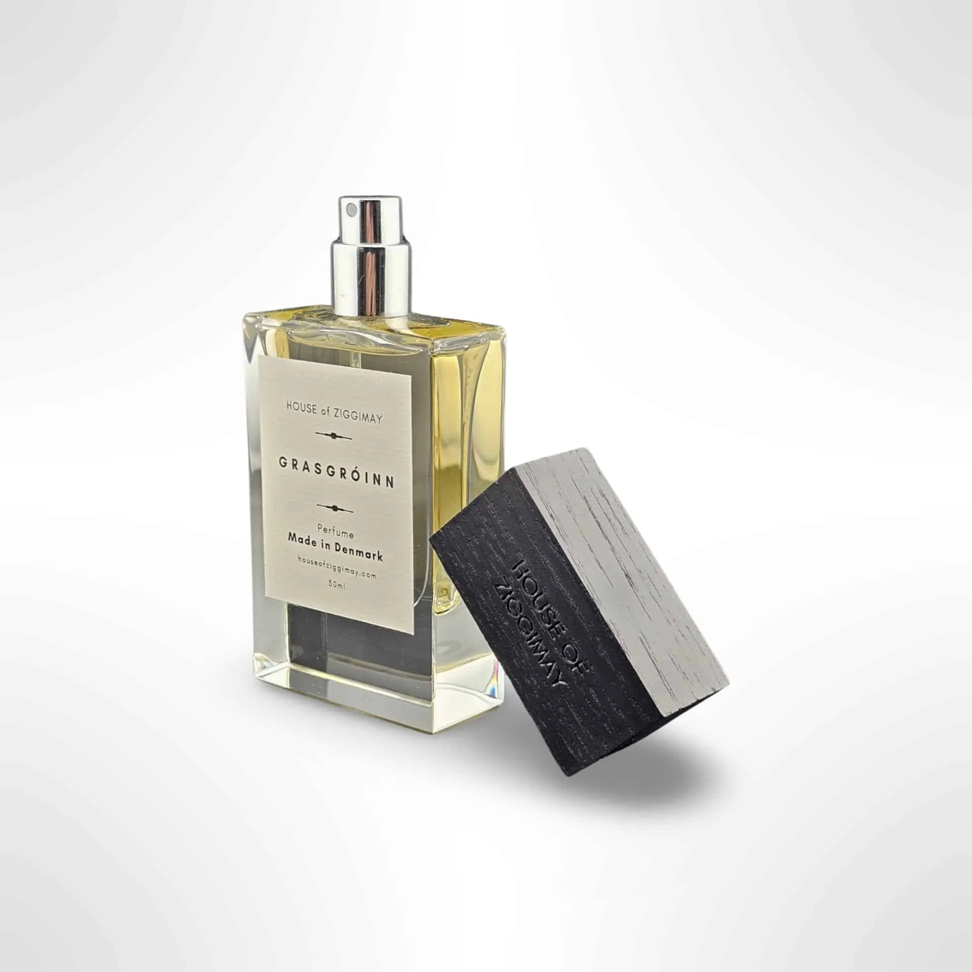 A glass perfume bottle featuring a metallic spray top and labeled Hous of Zigmay, Grasgróinn contains a yellow fragrance. A separate black cap is positioned nearby, and the label proudly indicates it is made in Denmark.