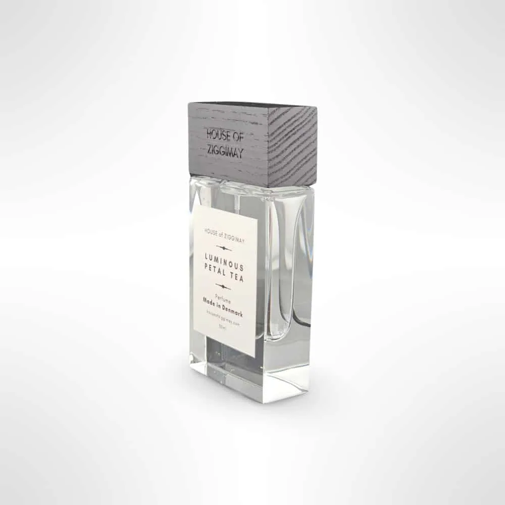 A sleek perfume bottle with a wooden top labeled HOUSE of ZIGGIMAY. The clear glass bottle reveals a pale yellow liquid inside. The front label reads HOUSE of ZIGGIMAY, Luminous Petal Tea, Eau de Parfum, Made in Denmark. This niche perfume stands against a solid white background.