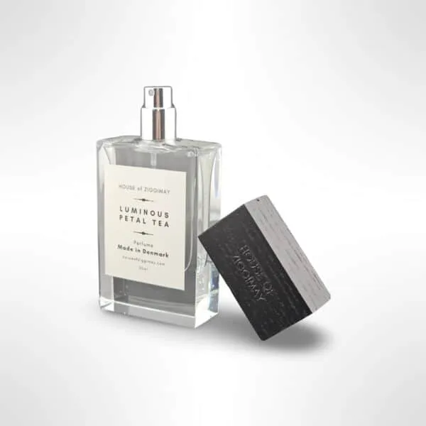 A clear rectangular bottle labeled Luminous Petal Tea from HOUSE of ZIGGIMAY emits a captivating fragrance. The bottle features a silver spray nozzle, and the black rectangular cap is placed nearby. This exquisite scent is masterfully crafted in Denmark.