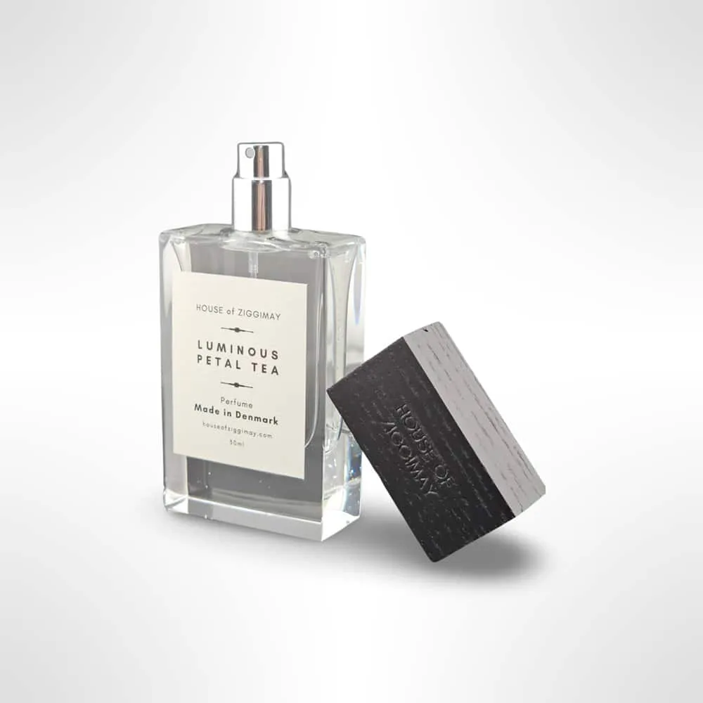 A clear rectangular bottle labeled Luminous Petal Tea from HOUSE of ZIGGIMAY emits a captivating fragrance. The bottle features a silver spray nozzle, and the black rectangular cap is placed nearby. This exquisite scent is masterfully crafted in Denmark.
