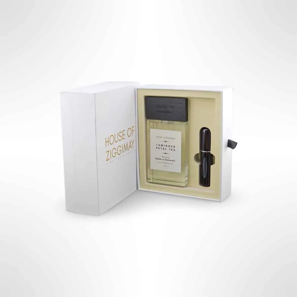 Perfume set showcasing a rectangular bottle of Luminous Petal Tea by HOUSE OF ZIGGIMAY, packaged elegantly in a white box adorned with gold lettering. This exquisite fragrance set includes a black cap and a small applicator, with the box partially open to reveal the captivating scent inside.