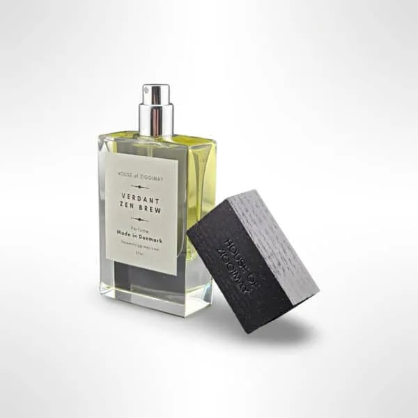 A bottle of Verdant Zen Brew, a niche perfume by HOUSE OF ZIGGIMAY, is featured. The rectangular glass bottle contains a light green liquid and showcases a label on the front. The black cap, removed, lies diagonally in the foreground against a plain gradient backdrop.