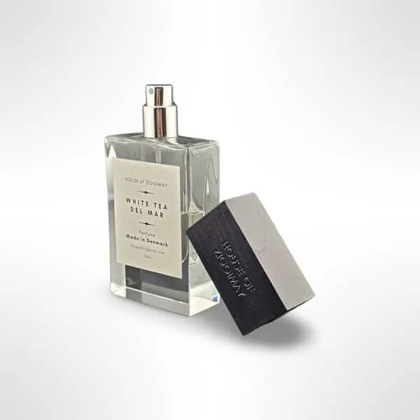 A perfume bottle and a black box. The bottle, with a silver cap, features a label that reads White Tea Del Mar, Perfume, Made in Denmark, 30ml. The black box beside it is embossed with the brand name HOUSE of ZIGGIMAY. The background is plain white. A touch of jasmine enhances the captivating fragrance of White Tea Del Mar.