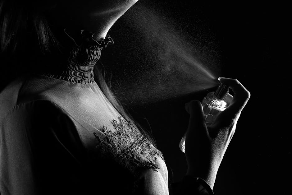A person in a lace-collared top sprays haute parfum onto their neck. The black and white image captures the silent luxury of the moment, with a delicate mist from the perfume visible in the light.