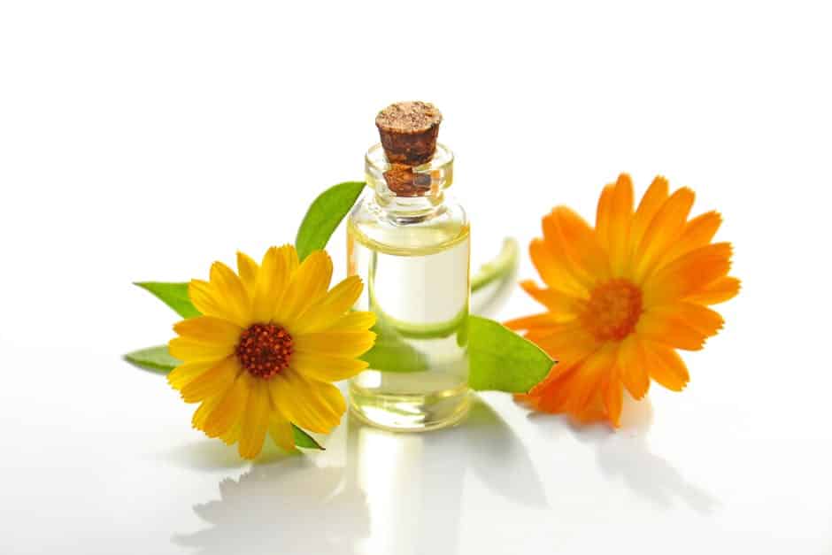 A small glass bottle of clear Eau de Parfum with a cork stopper is placed between two bright yellow and orange flowers with green leaves on a white background.