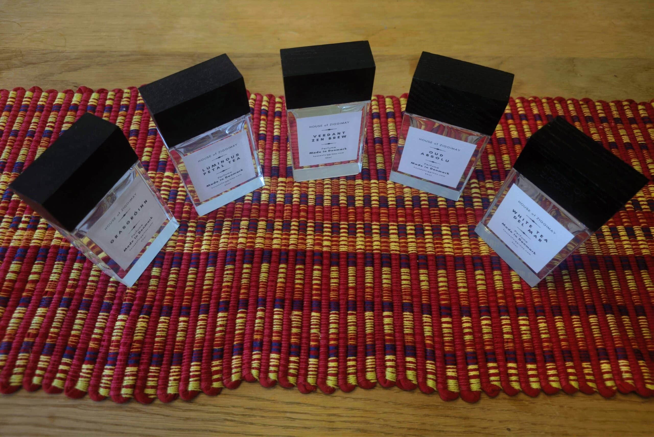 Five rectangular bottles of luxury perfumes with black caps are arranged in a semi-circle on a colorful woven red, yellow, and purple placemat, placed on a wooden table.