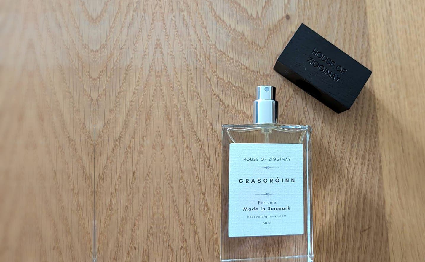 A Grasgróinn perfume bottle beside its ash wood cap on an oak table, the label reading HOUSE OF ZIGGIMAY, Perfume, Made in Denmark