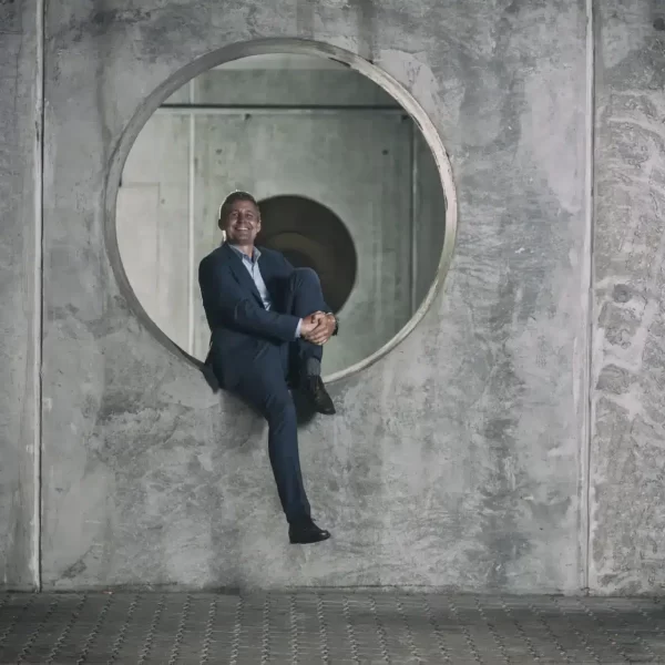 A person in a suit sits playfully within a circular cutout in a concrete wall. He is smiling and has one leg crossed over the other, dangling outside the circle. The industrial background features grey, textured concrete, subtly echoing the sophisticated craftsmanship of HOUSE of ZIGGIMAY's niche perfume.