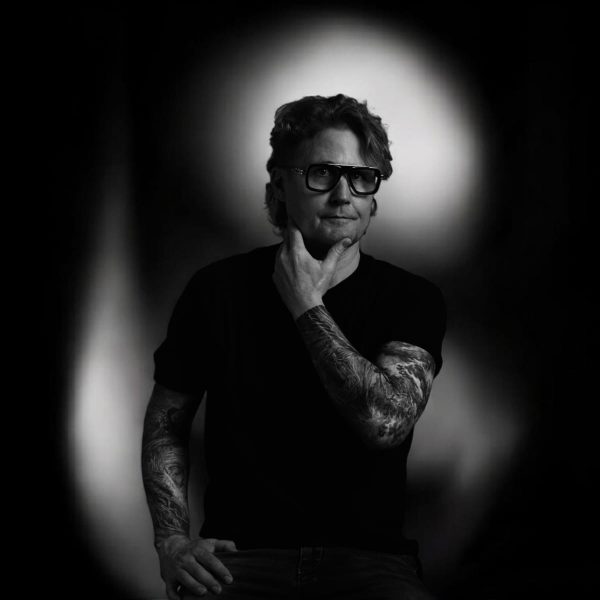 Black and white portrait of a person with tattoos, wearing glasses and a black shirt, posing with one hand on their chin. The background is softly lit, creating a halo effect around the figure.